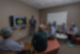Man presenting to a group wearing hard hats, training presentation on screen.